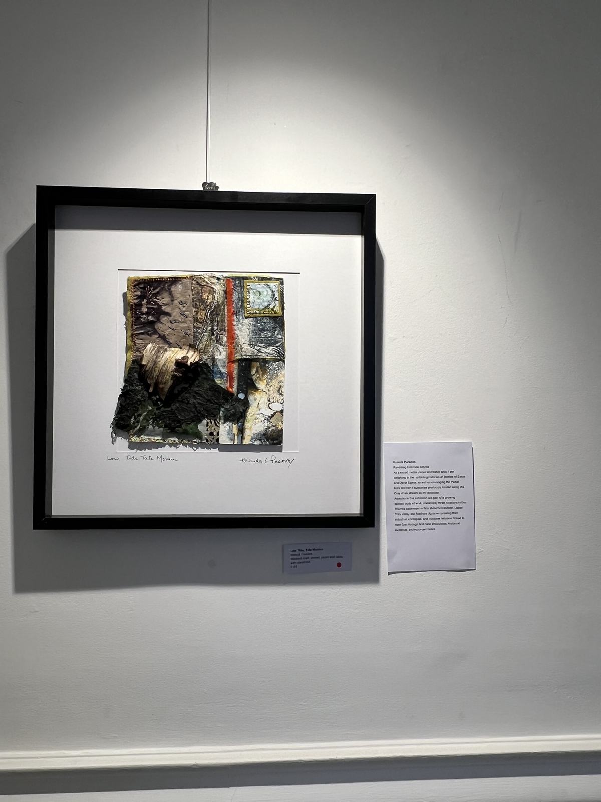 Dark gallery installation showing a framed mixed media work under a pool of light